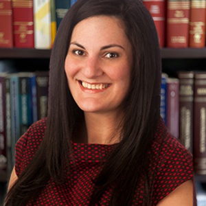 Ariane Raneri, a woman with long dark hair wearing a red and black patterned top, smiles in front of a bookshelf filled with books.