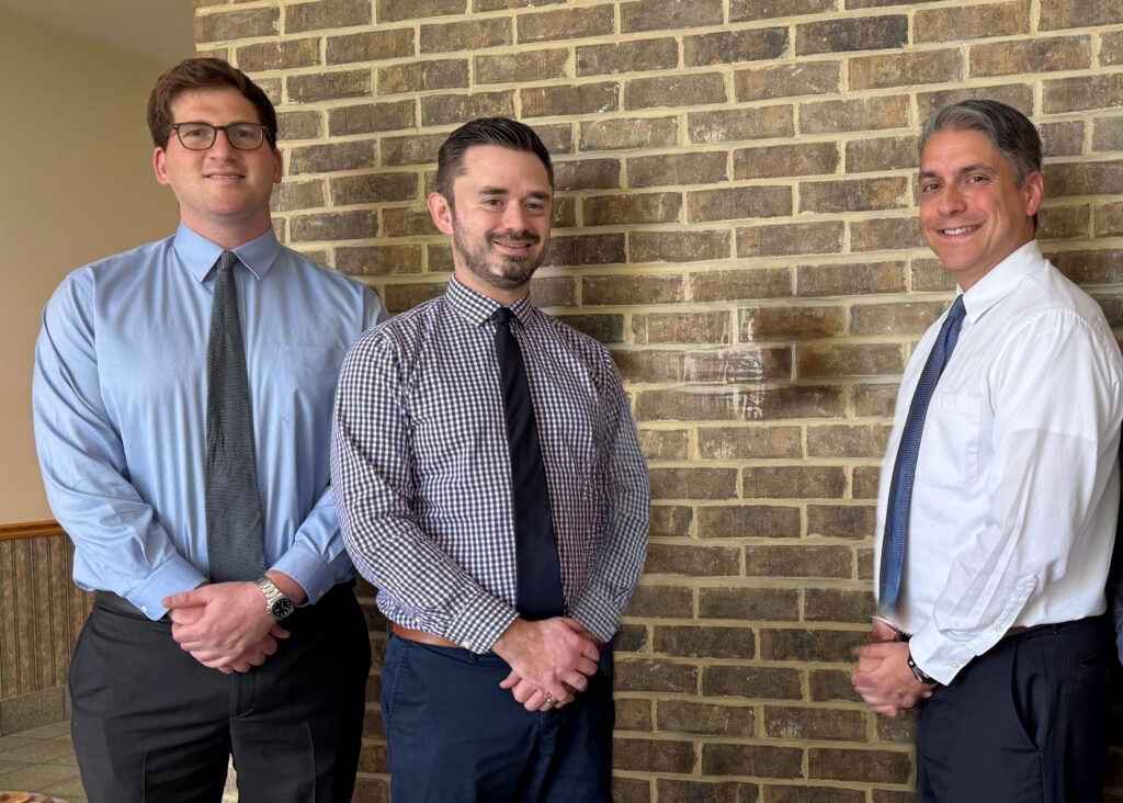 Three men in button-down shirts and ties stand side by side in front of a brick wall, smiling at the camera with their hands folded in front of them—an image that perfectly captures the camaraderie and professionalism at the heart of our About Us story.