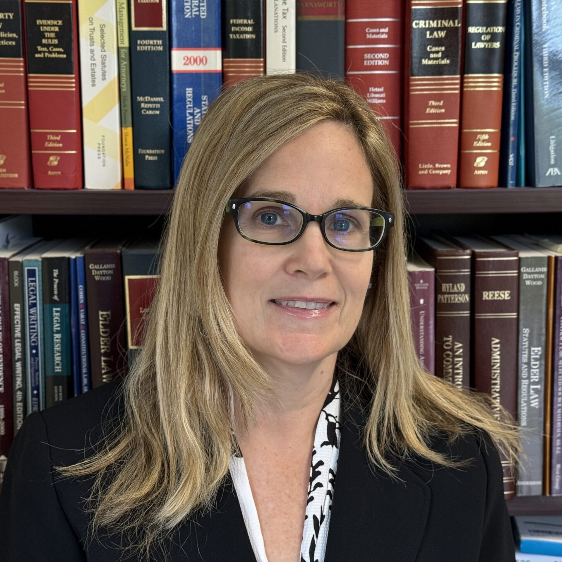 A woman with long blonde hair and glasses wearing a black blazer, posed in front of a bookshelf filled with law textbooks.